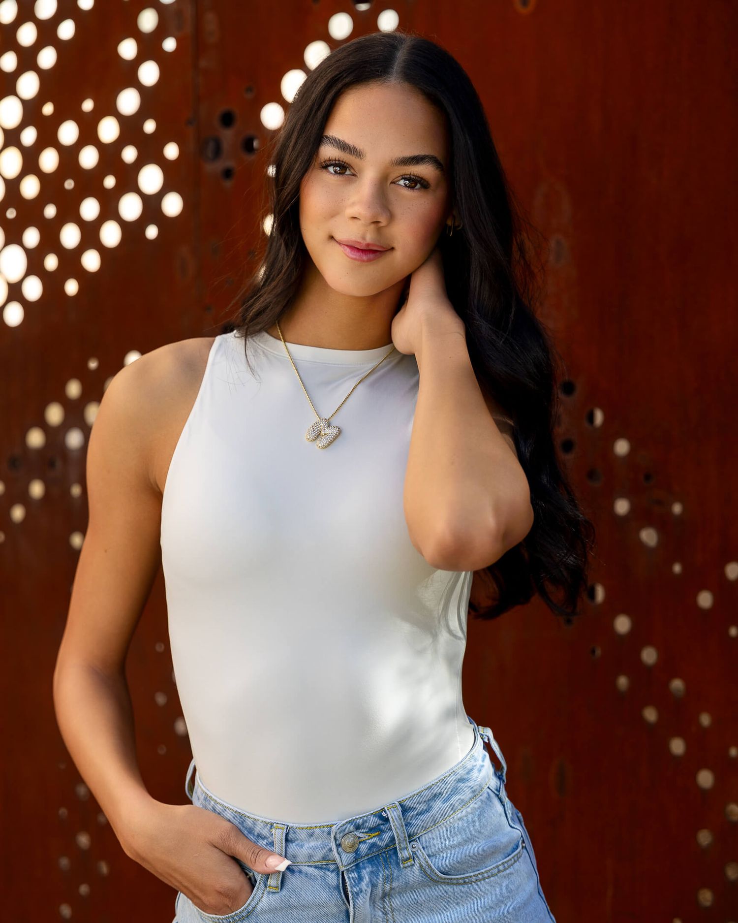 Young woman in white sleeveless top and jeans wearing gold necklace posing against rustic metal wall.