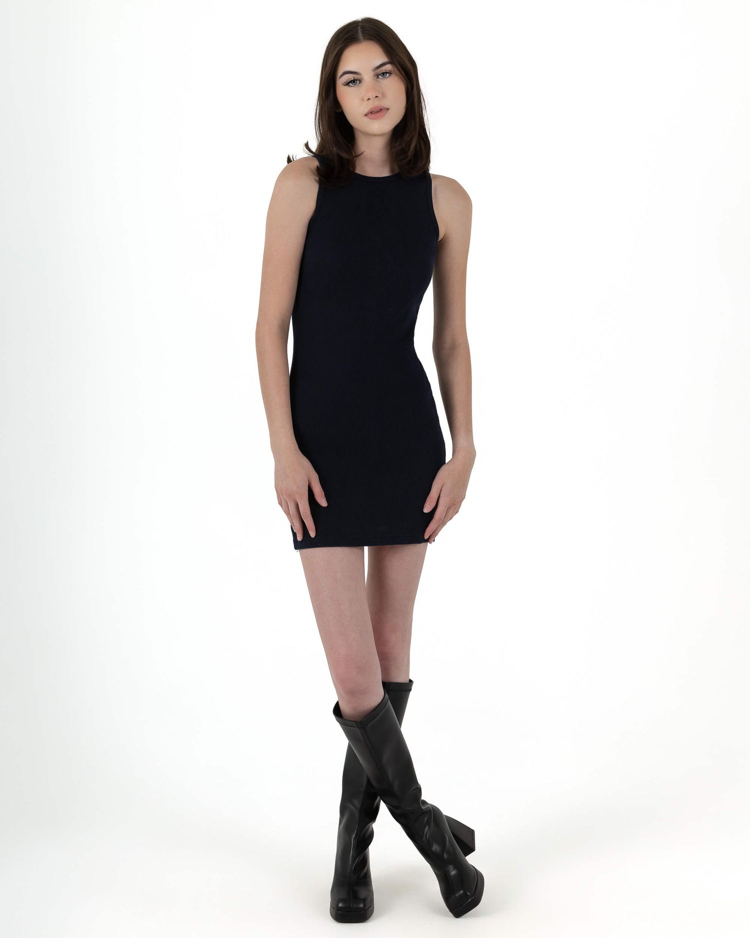 Woman in sleeveless black mini dress with knee-high boots posing on white background.