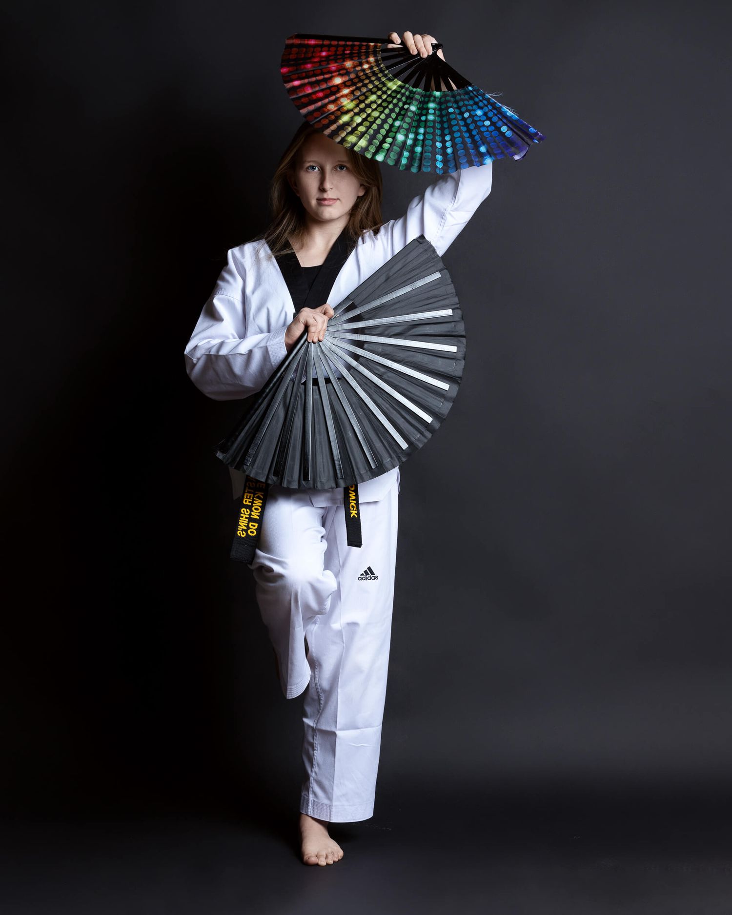 Young taekwondo athlete in white uniform poses with colorful fans on dark background.