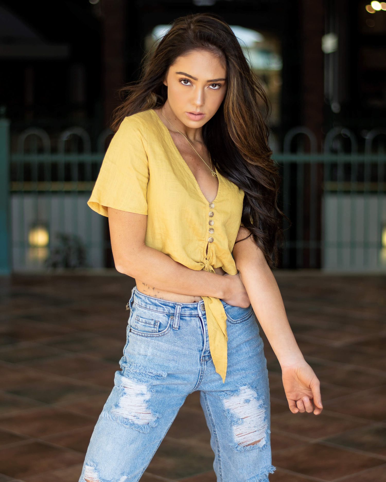 Young woman in yellow crop top and ripped jeans posing indoors.