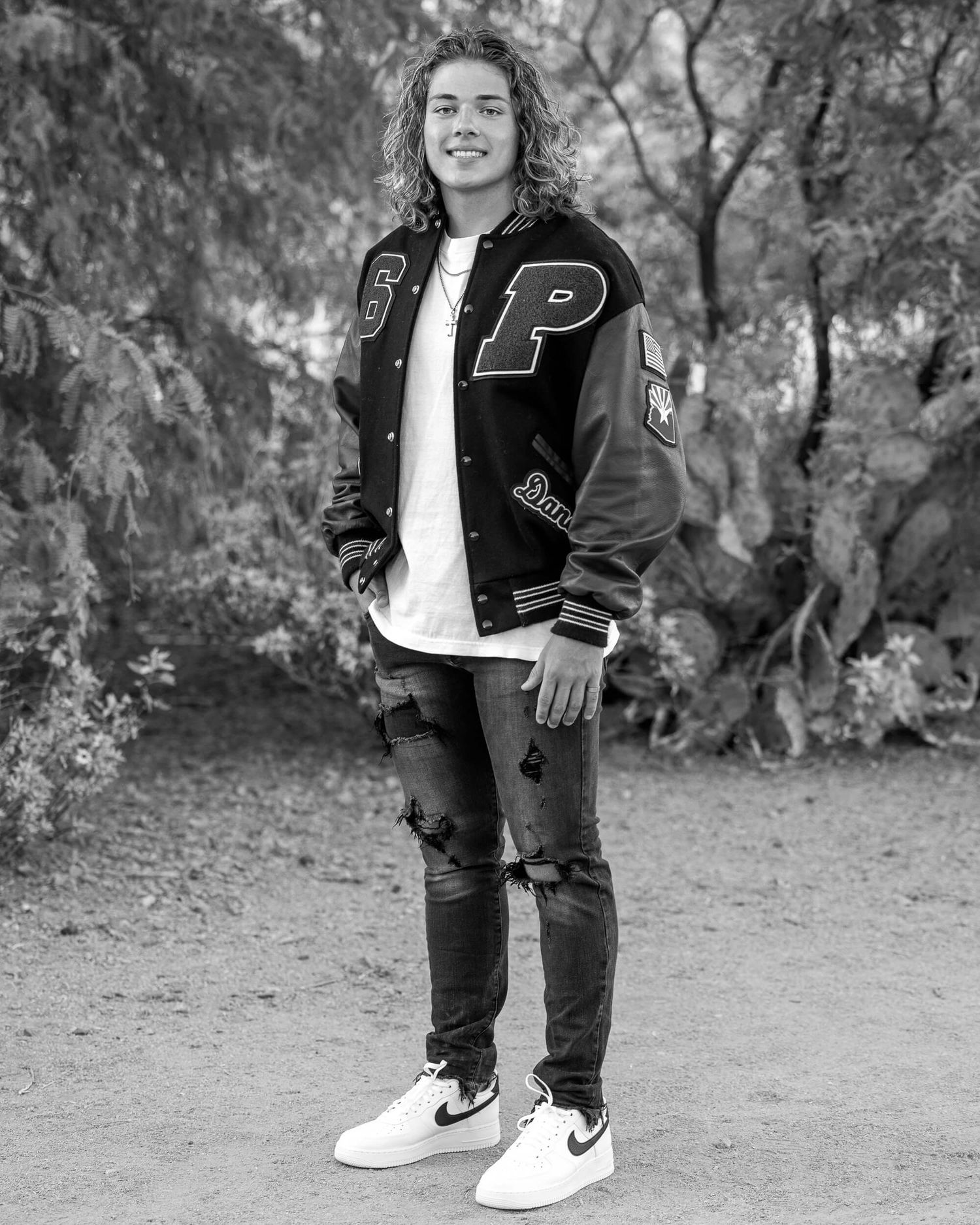 Young man in letterman jacket, ripped jeans, and white sneakers posing outdoors in black and white photo.