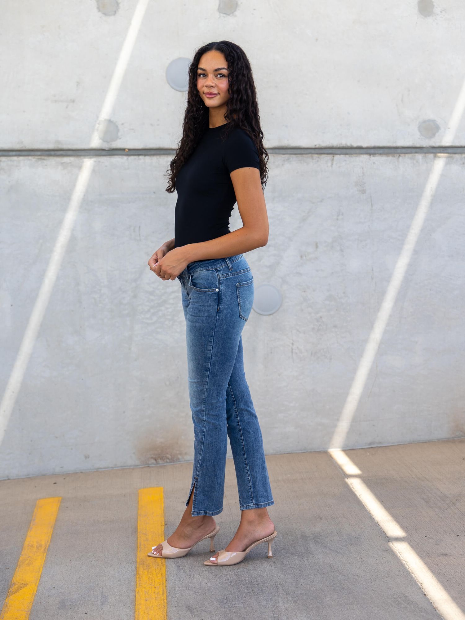 Woman in black tee, blue jeans, and pink heels posing sideways against a concrete wall outdoors.