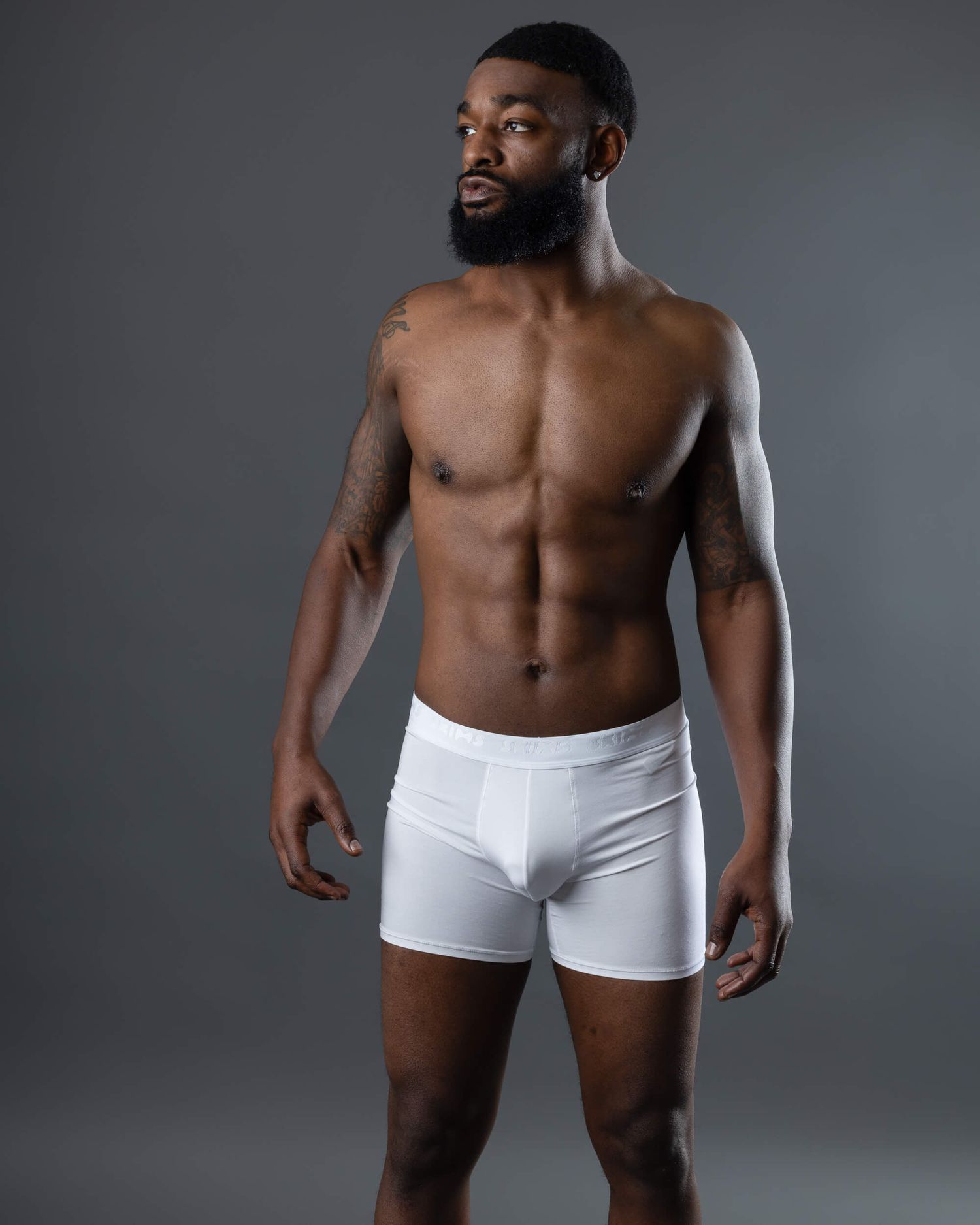 Muscular man with beard wearing white boxer briefs, posing confidently against gray background.