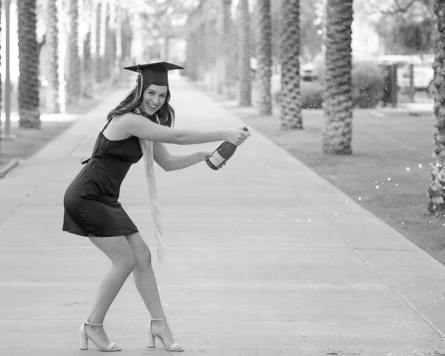 Black and white photo of graduate in black dress and cap playfully posing on palm tree-lined campus path.