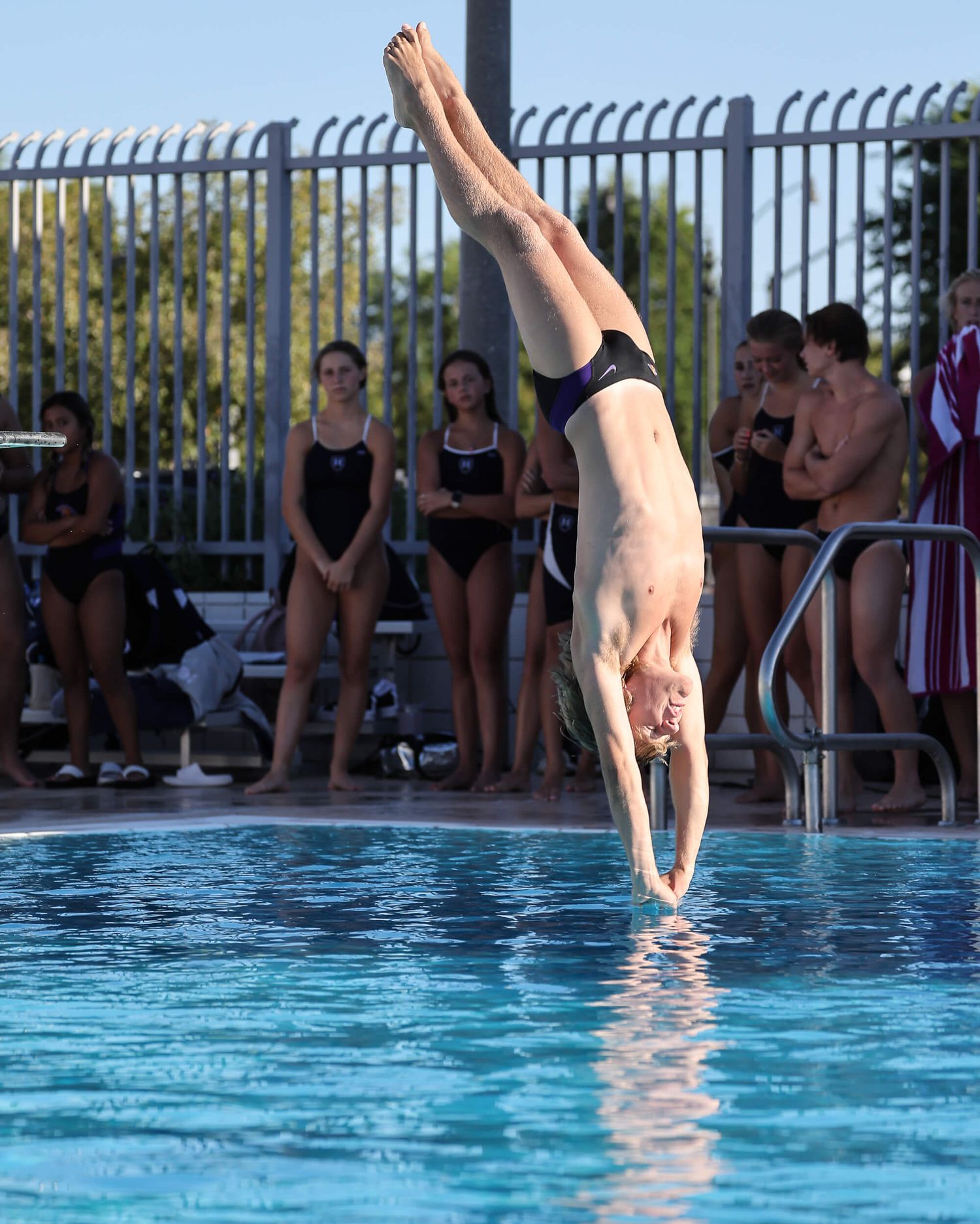 Male diver performs vertical entry dive into outdoor swimming pool before spectators.