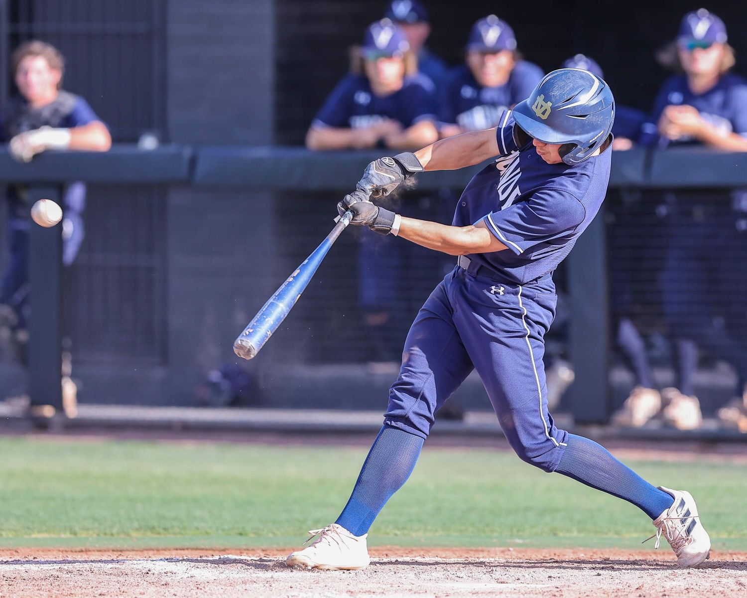 Baseball player in navy uniform swings bat during a college game with teammates visible in the dugout background.