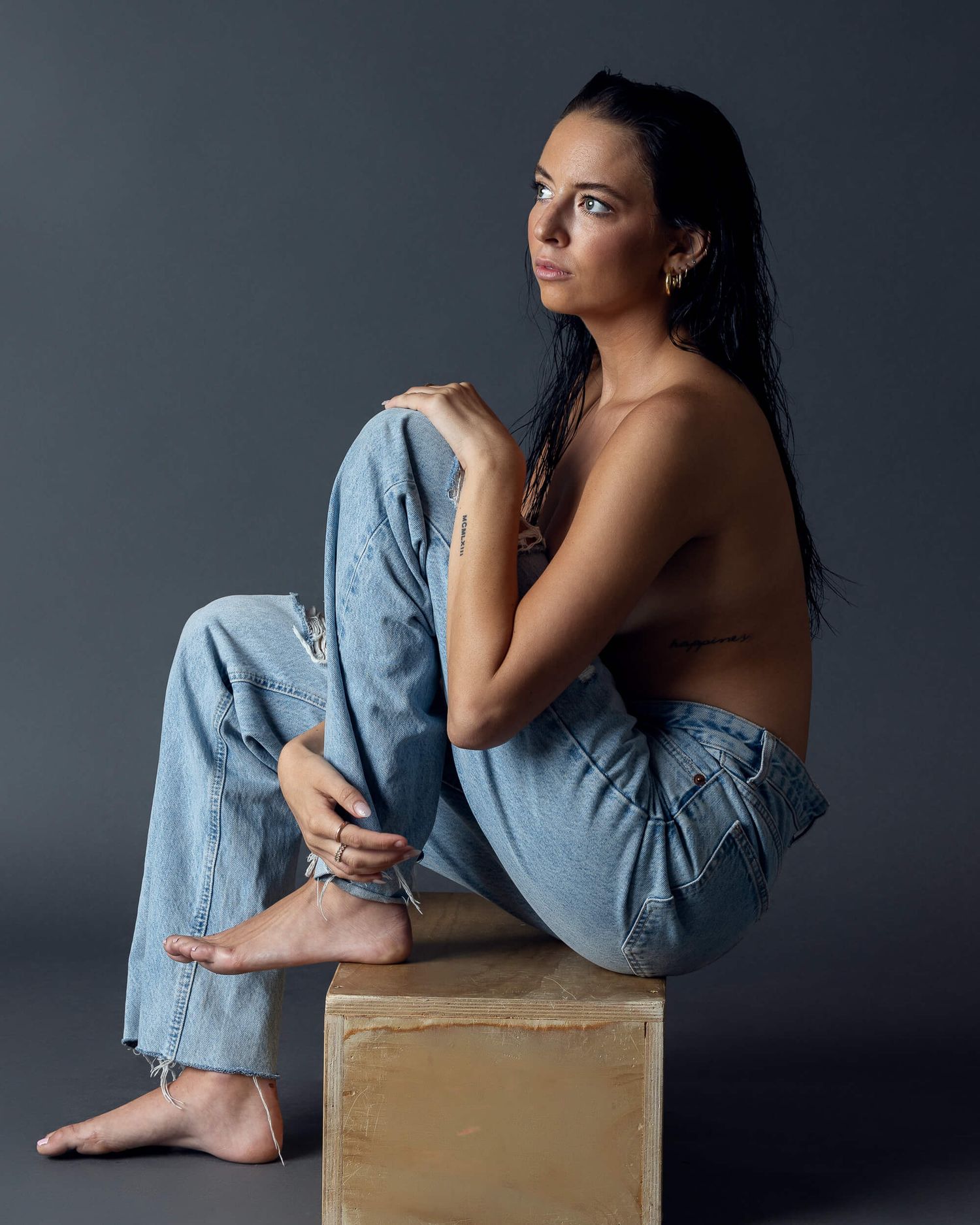 Brunette woman sits topless in jeans on wooden block in artistic studio portrait.