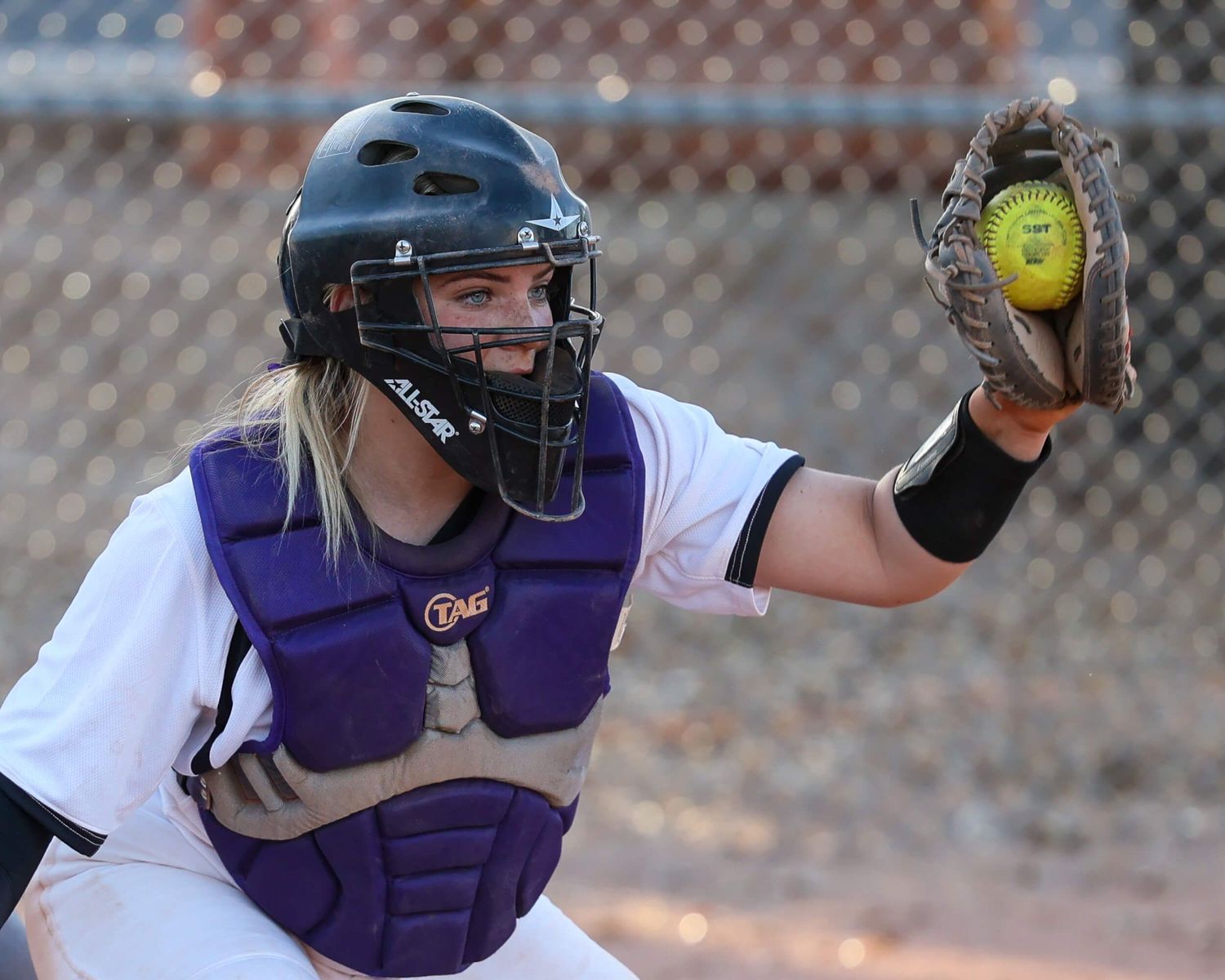 Female softball catcher in purple gear catches a yellow ball during a game.