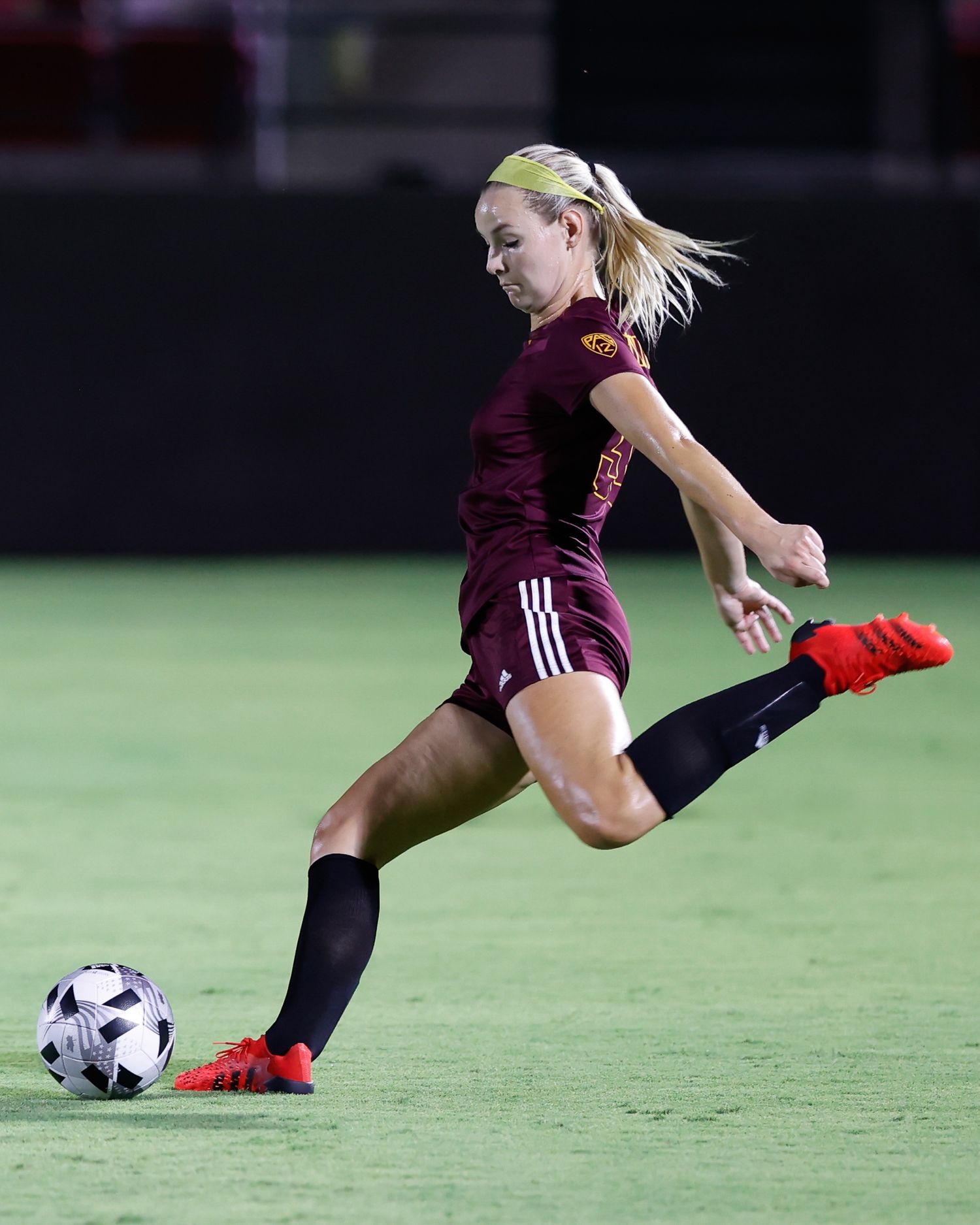 Female soccer player in maroon uniform kicking ball during night game on illuminated field.