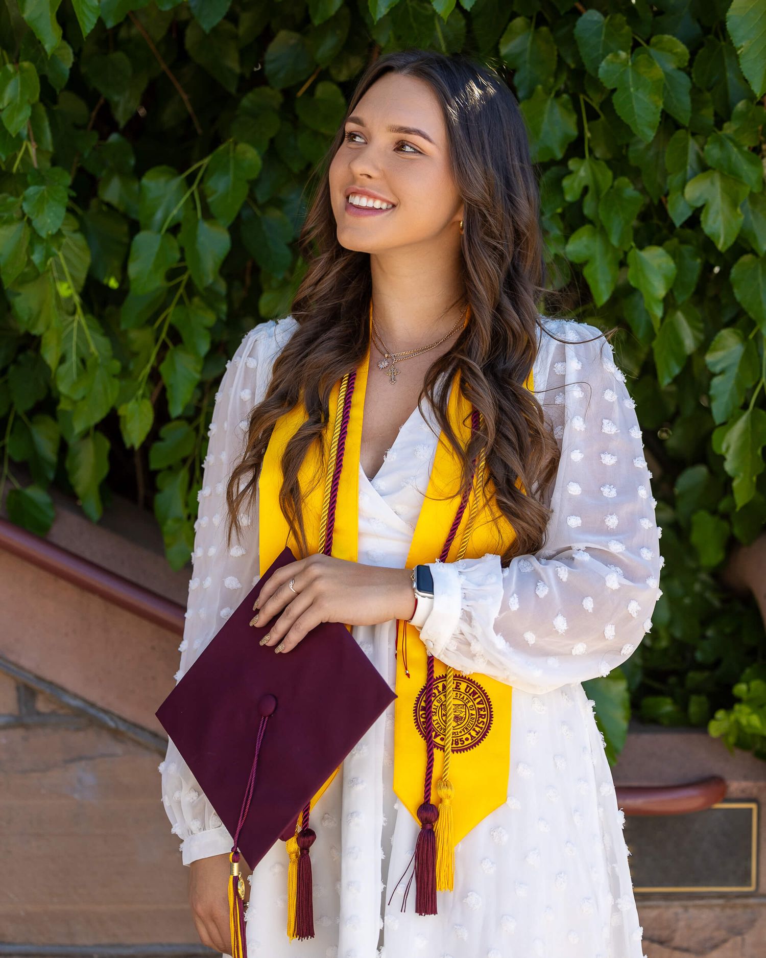 Smiling graduate in white dress holding maroon cap with gold ASU stole outdoors.