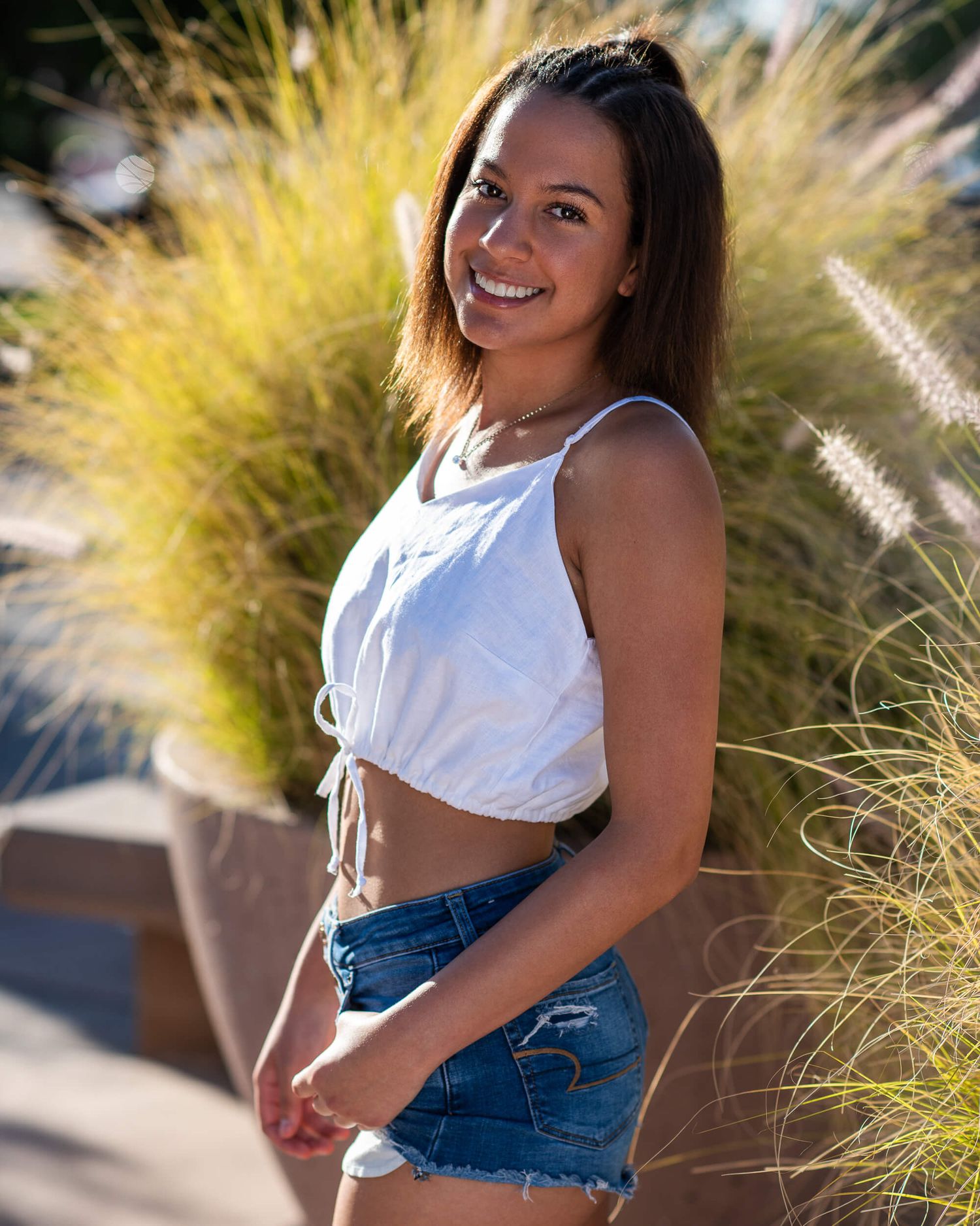 Smiling young woman in white crop top and denim shorts posing outdoors near ornamental grass.