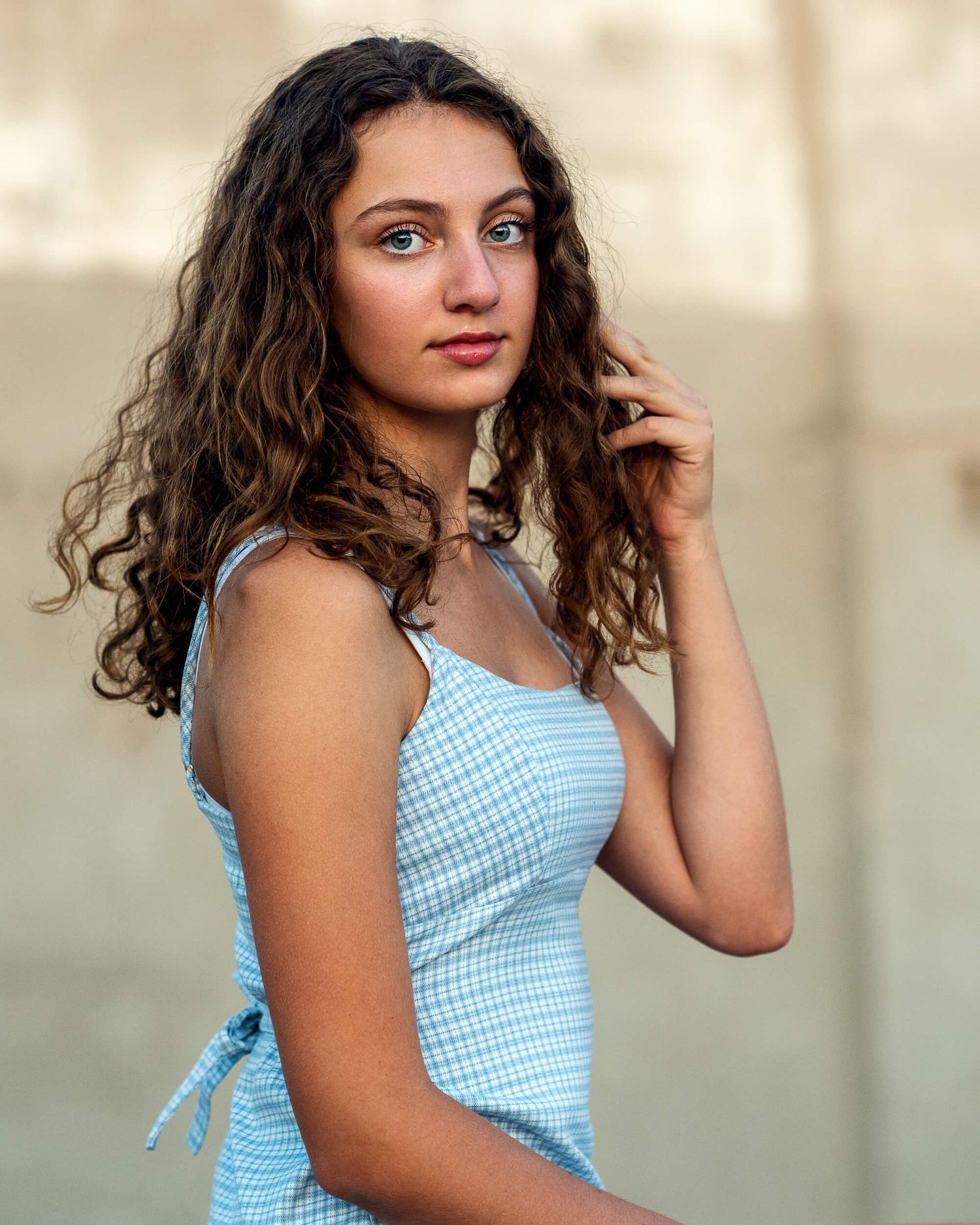 Young woman with curly brown hair wearing a light blue gingham dress, posing outdoors with a soft blurred background.