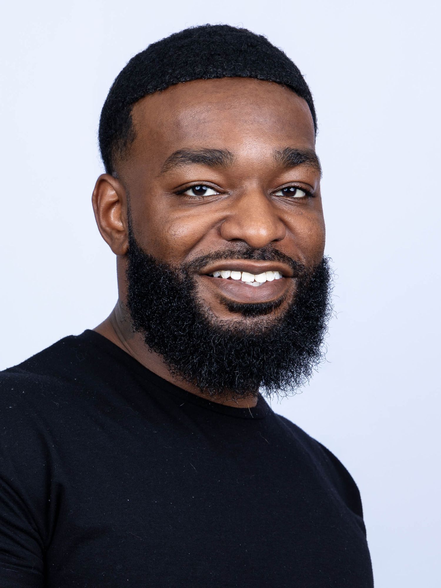 Smiling Black man with a full beard wearing a black t-shirt against a light gray background.