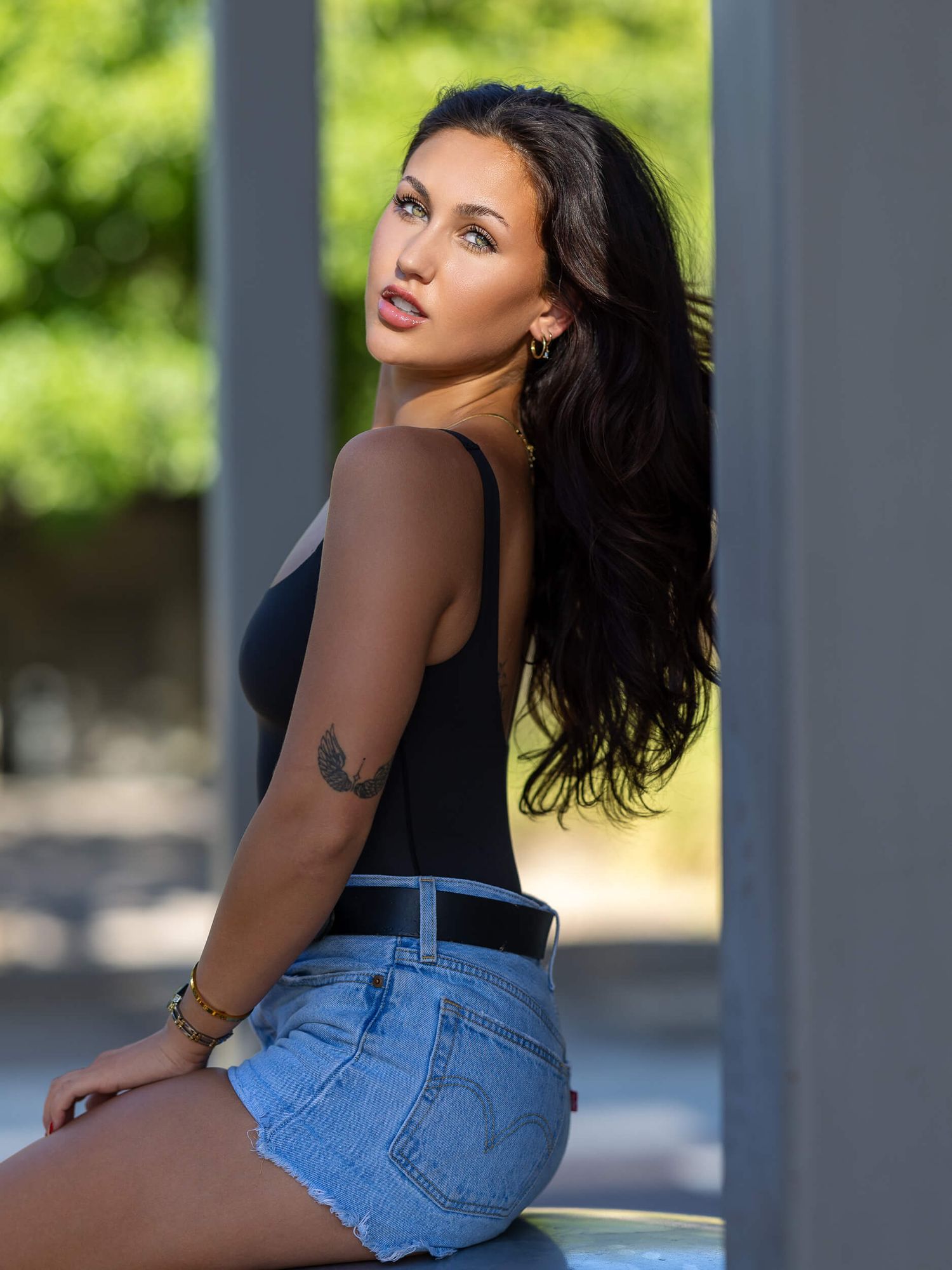 Young woman with long dark hair poses in black bodysuit and denim shorts, showcasing arm tattoo in sunny outdoor setting.