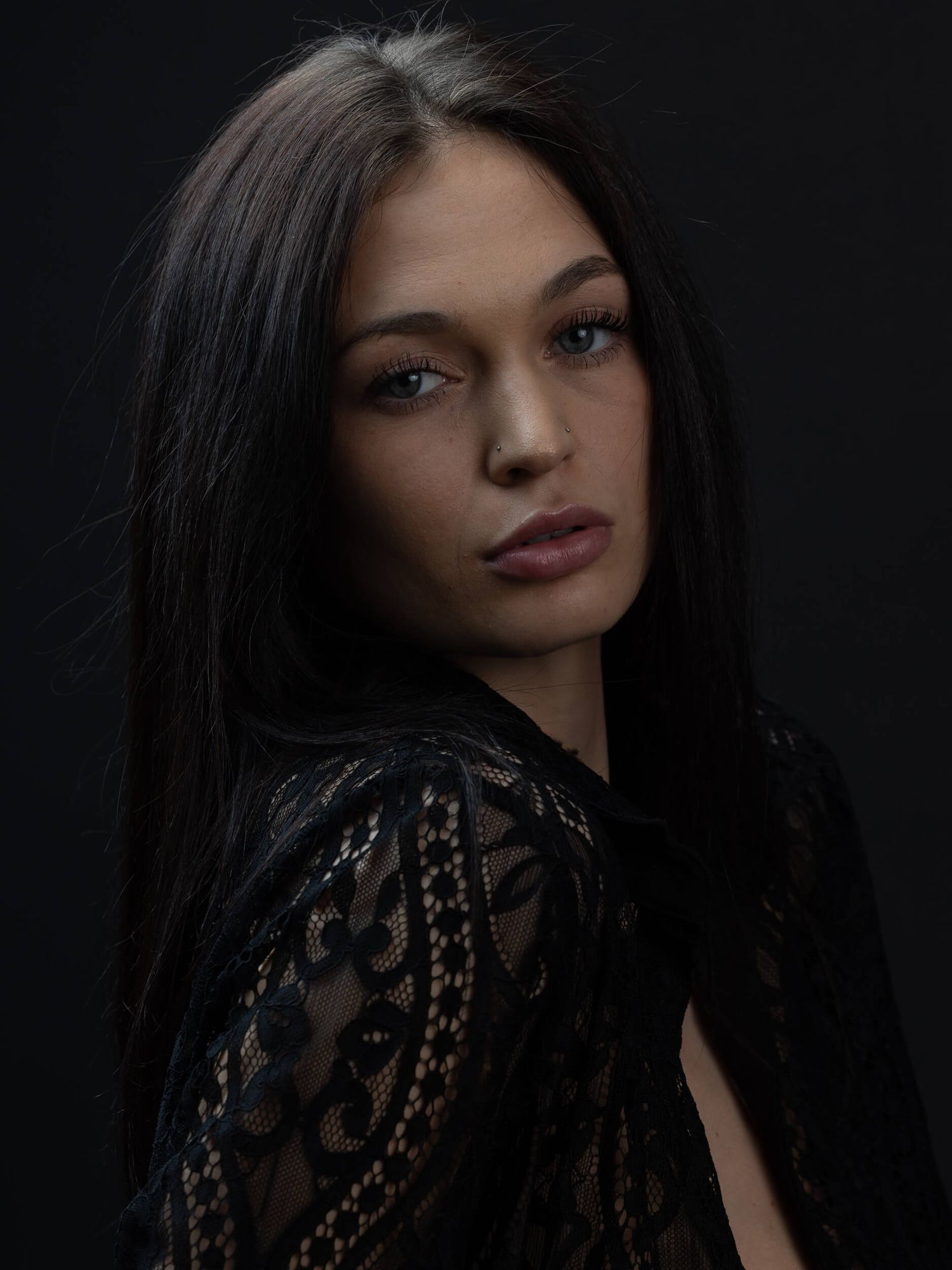 Young woman with long dark hair wearing black lace top, posing against dark background in moody portrait.