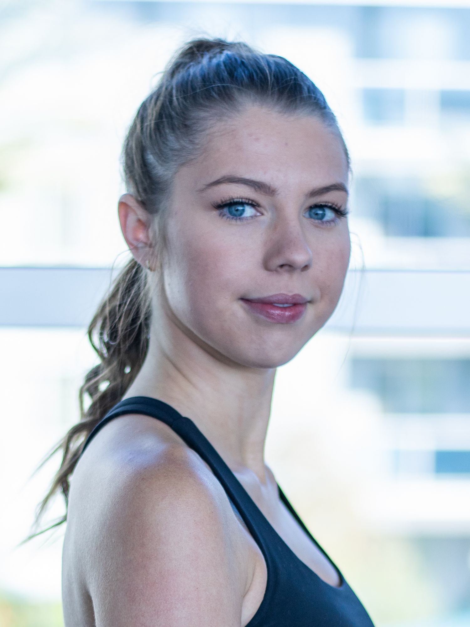Young woman with blue eyes and ponytail wearing black tank top poses confidently indoors near bright window.