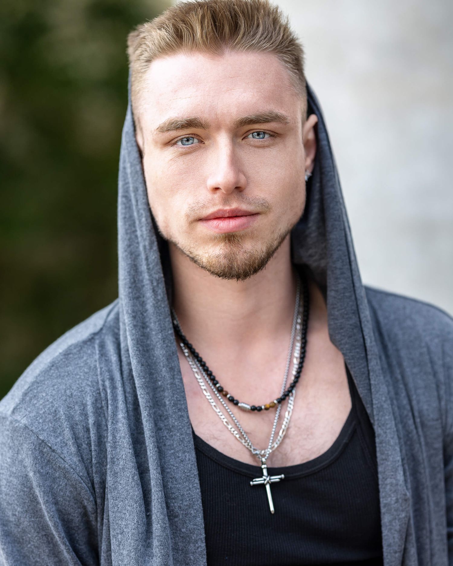 Blonde man with blue eyes wearing gray hoodie and cross necklace posing outdoors.