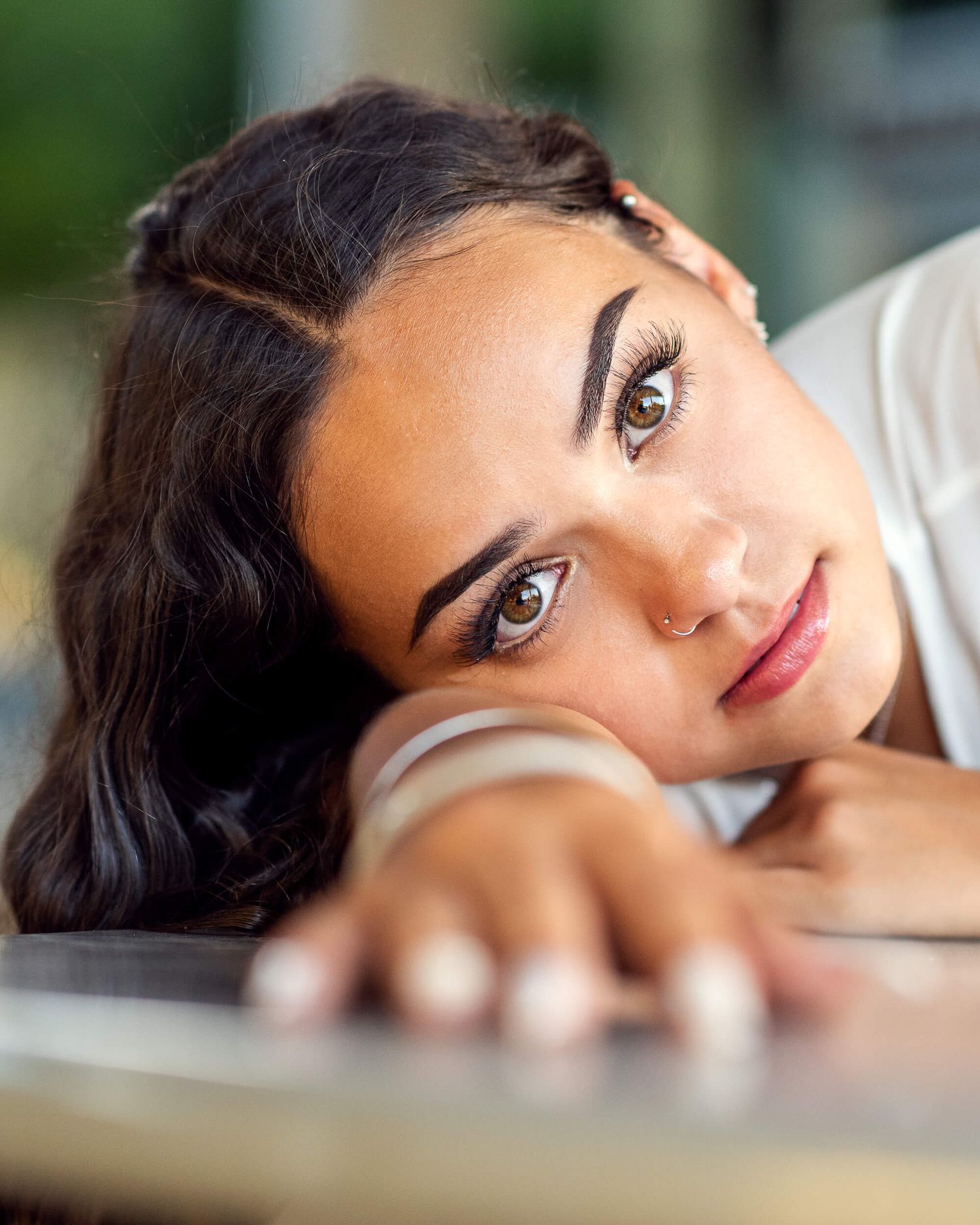 Dark-haired young woman with nose ring resting chin on hands gazing at camera indoors.