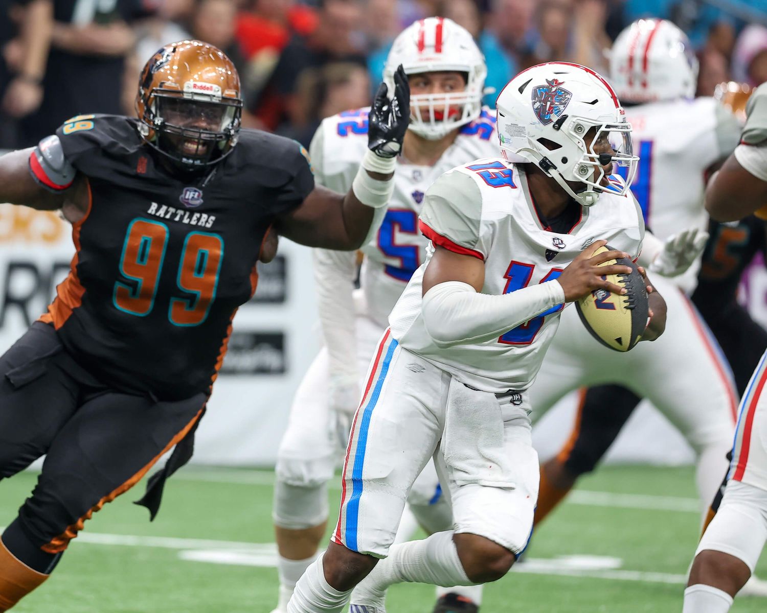Football player wearing number 99 Rattlers jersey rushes quarterback during intense indoor arena football game action.