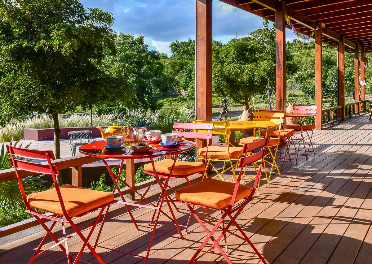 Colorful red and orange bistro tables and chairs on a sunny wooden deck surrounded by green trees.