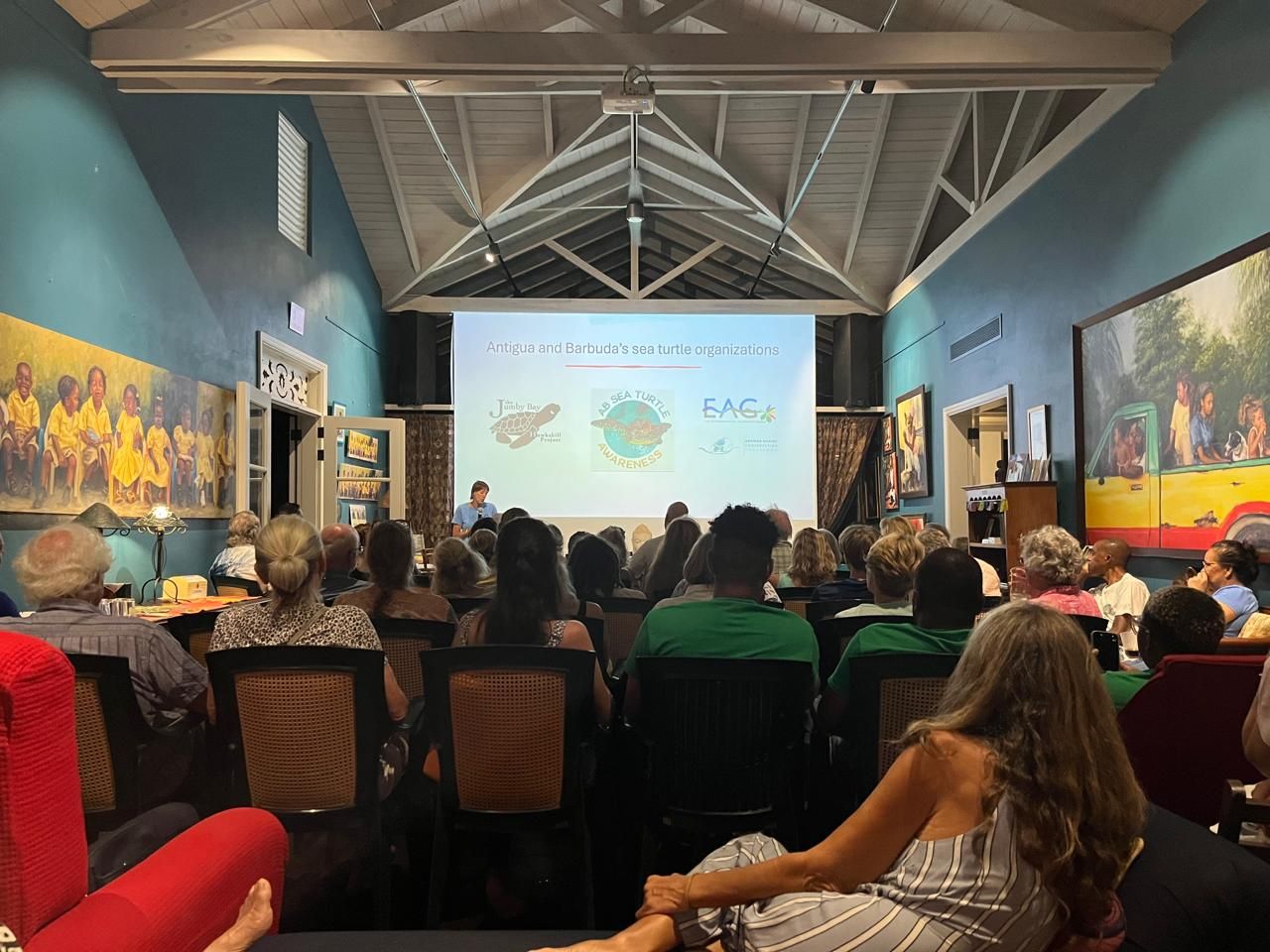 Audience attending a presentation in a colorful art-decorated hall with a projected screen displaying logos.