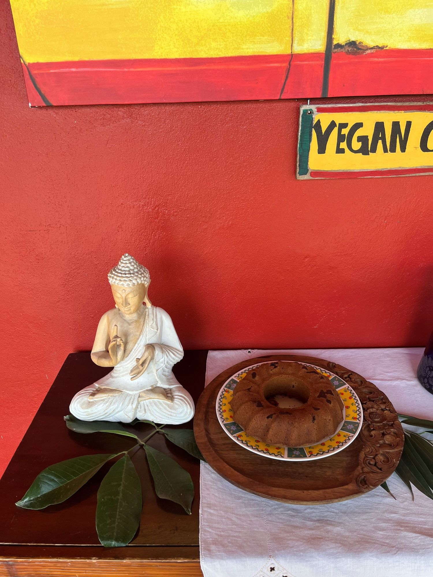 White Buddha statue beside a chocolate bundt cake on a wooden table at a vegan restaurant with red walls.