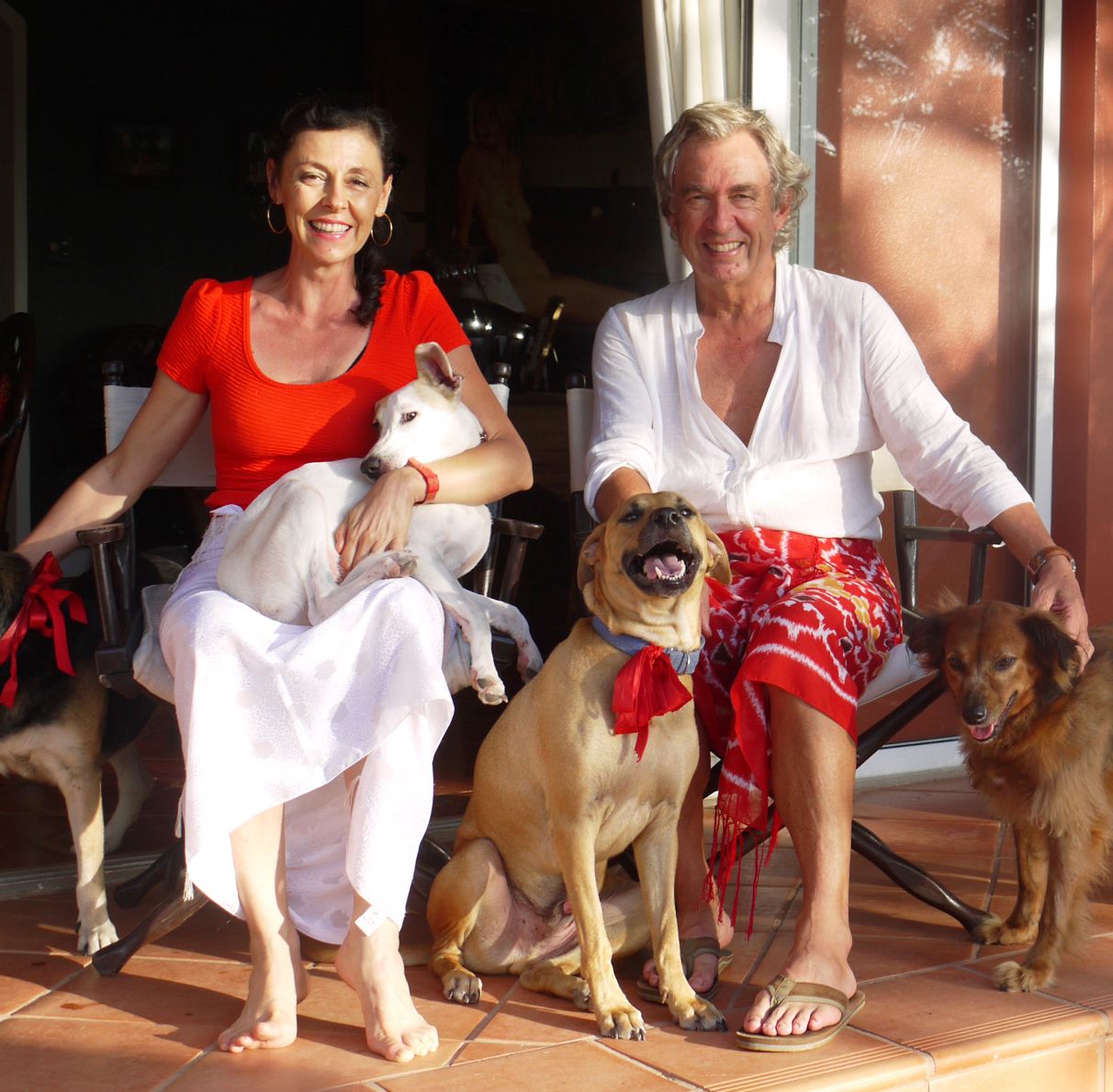 A smiling couple sits outdoors with four dogs, woman in red top holding white dog, man in white shirt with red shorts.