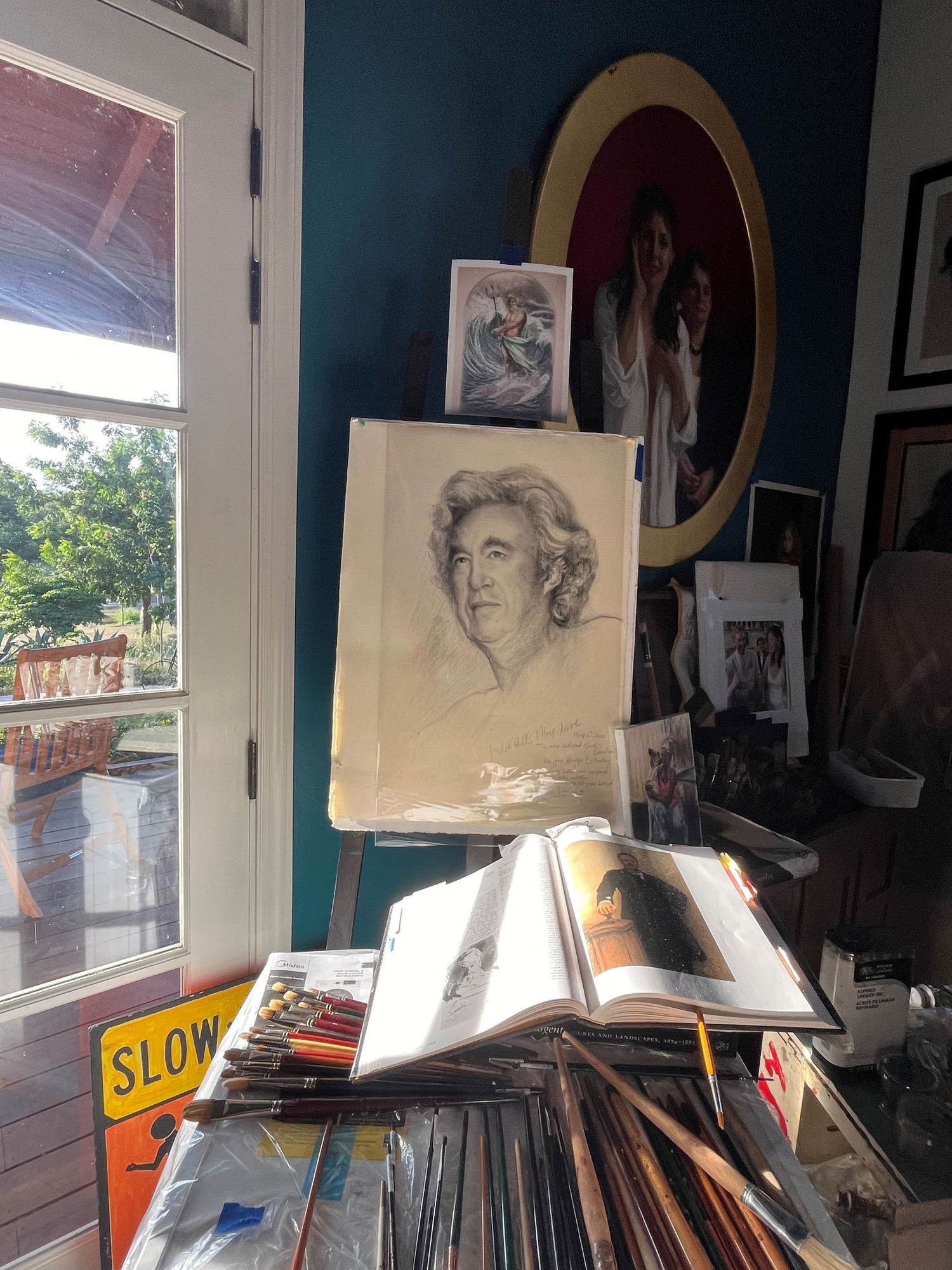 Artist studio with pencil portrait on easel, art books, brushes, and framed photos on teal wall.