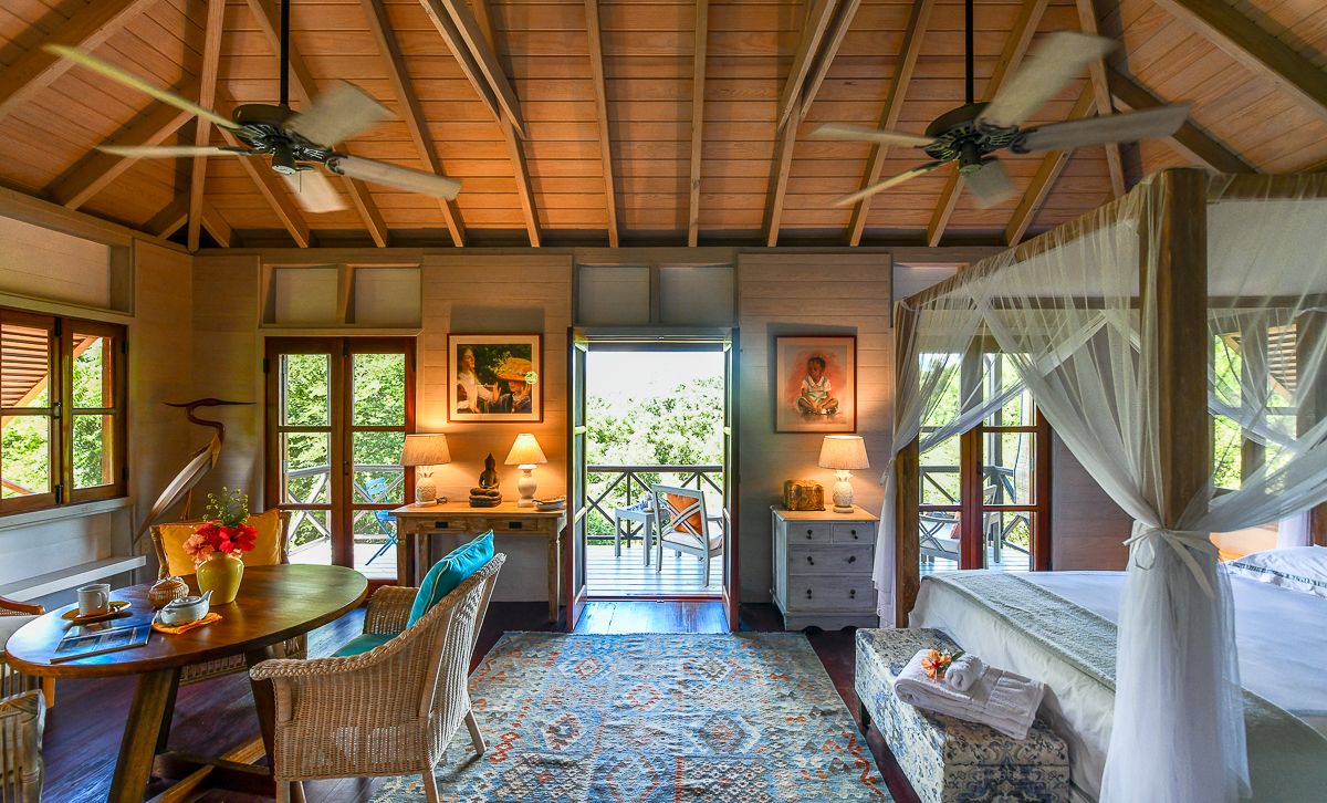 Elegant tropical cottage bedroom with canopy bed, ceiling fans, wicker furniture, and garden views.
