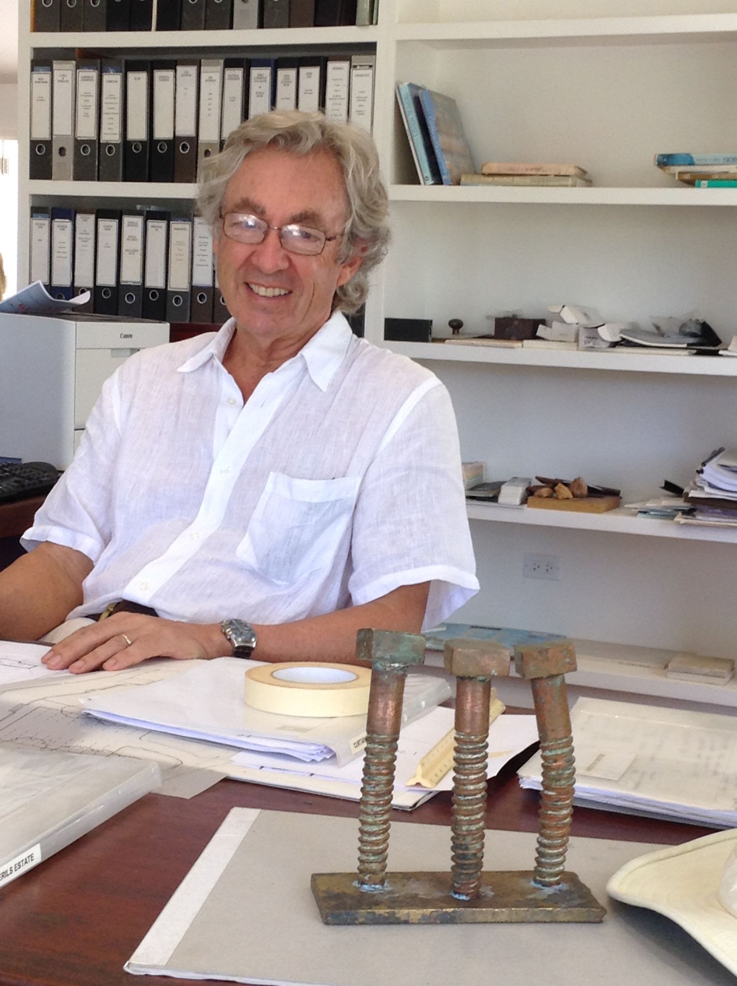 Smiling older man with gray hair and glasses sitting at a desk in a bright office with binders on shelves behind him.