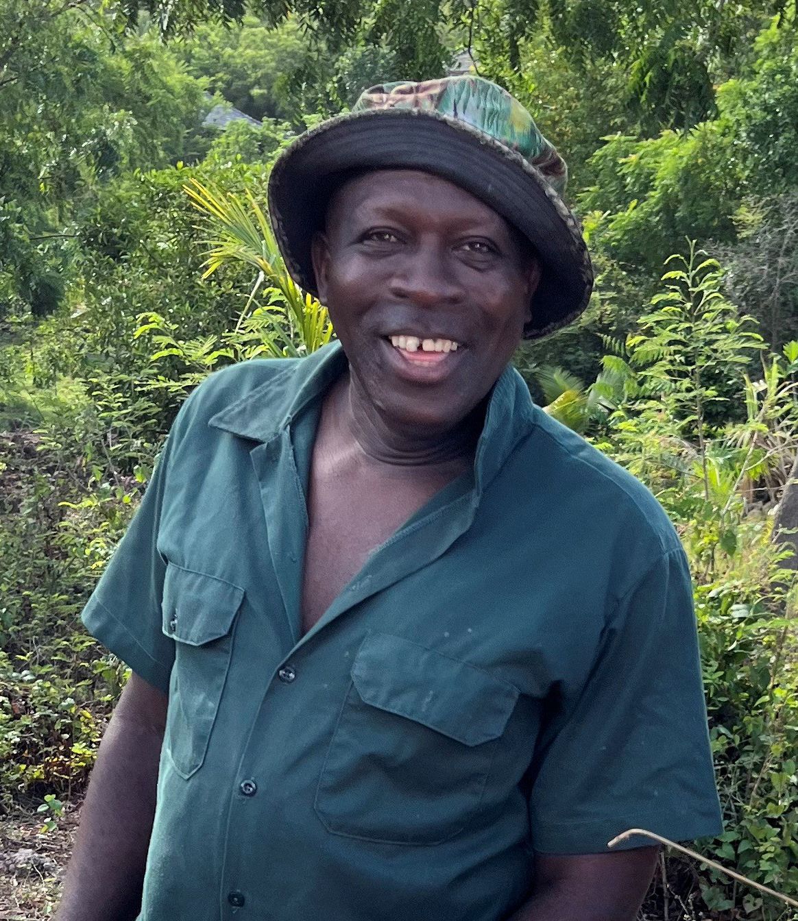Smiling man in green shirt and bucket hat standing outdoors surrounded by lush tropical greenery.