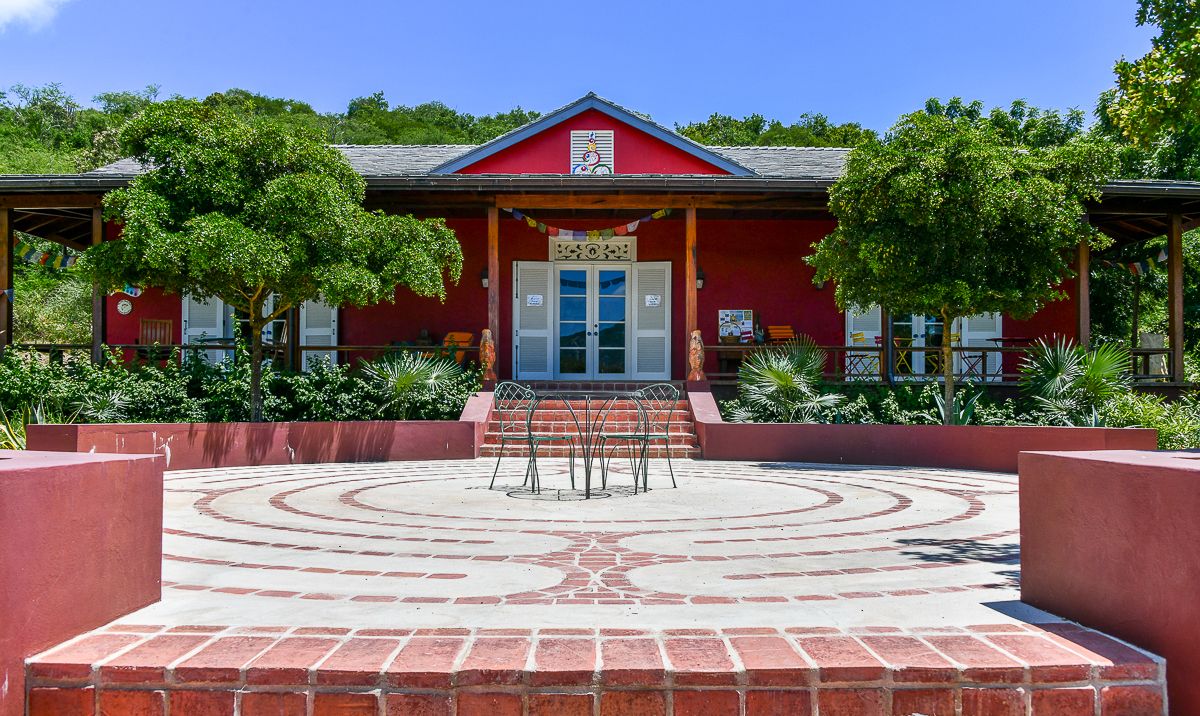 Red building with white doors, brick labyrinth courtyard, and lush green trees in a serene outdoor setting.