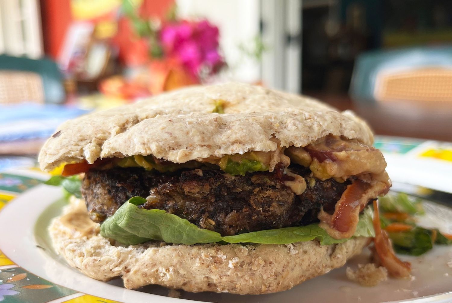 A hearty veggie burger with avocado, bacon, and fresh greens on a rustic bun served on a white plate.