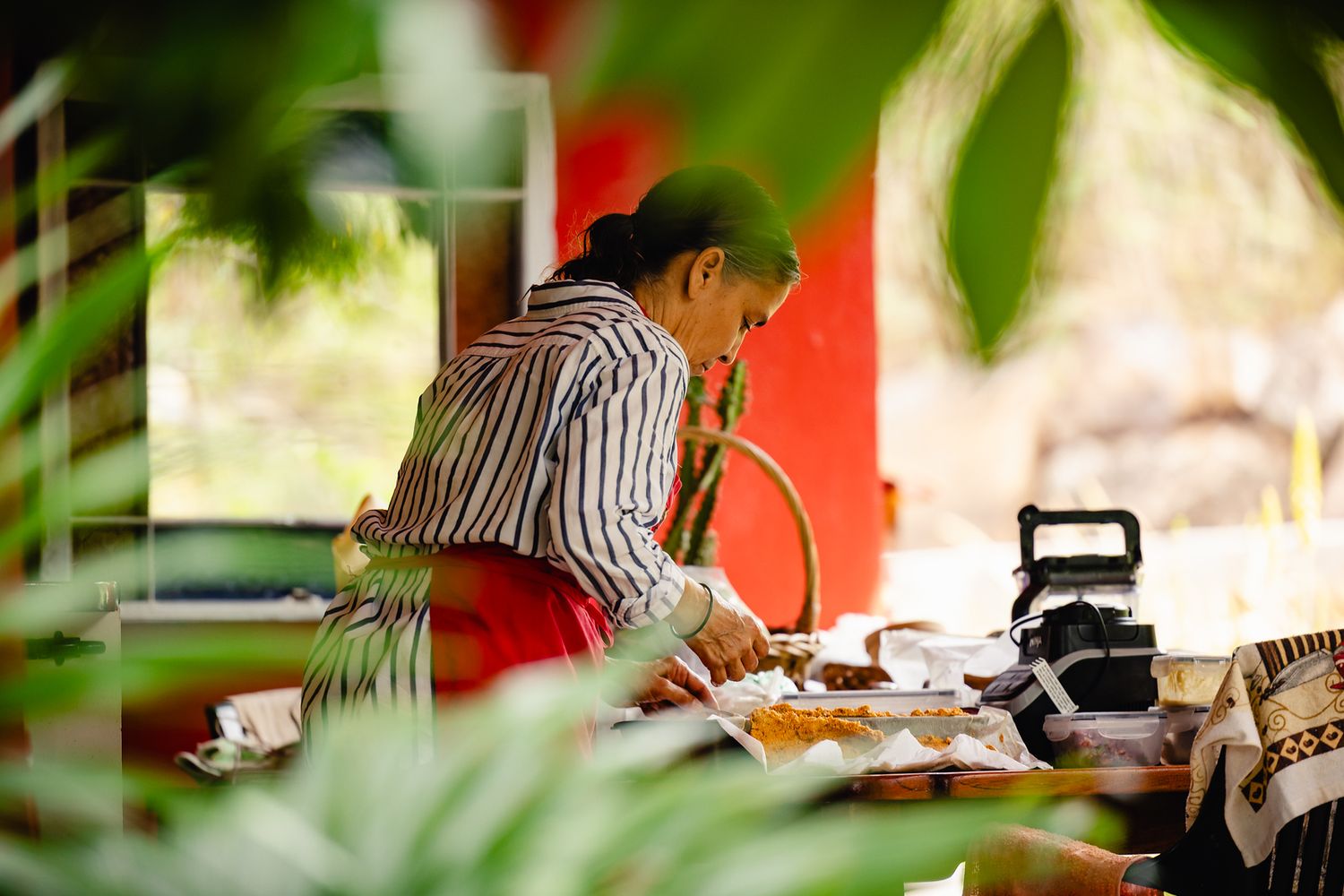 Woman in striped shirt preparing food at an outdoor table surrounded by lush green tropical plants.