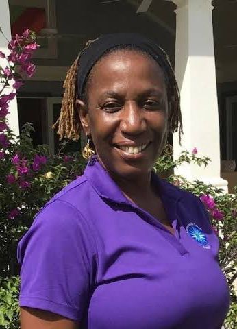 Smiling woman wearing a purple polo shirt and black headband, standing outdoors near colorful pink flowers.