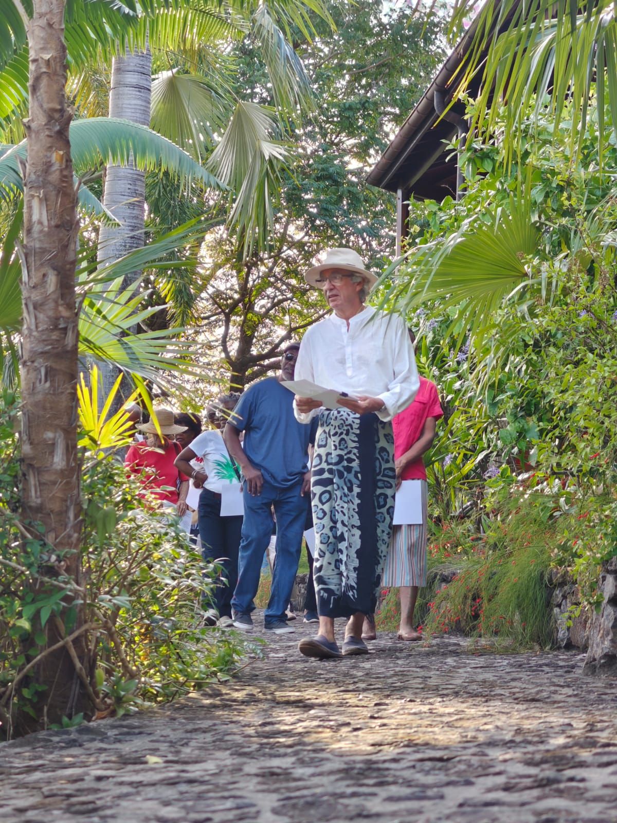 Elderly man in white shirt and straw hat walks cobblestone path through lush tropical garden with palm trees.