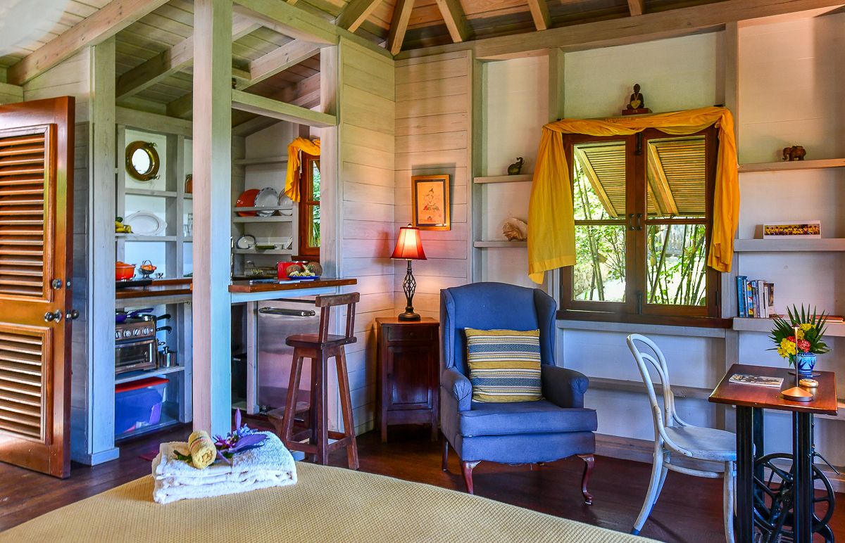 Cozy tropical cottage interior with blue armchair, yellow curtains, wooden walls, and open kitchen area.