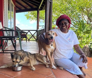 A smiling woman in a red hat and white outfit sits on a porch with two dogs on terracotta tiles.