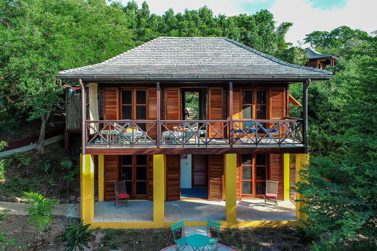 Two-story tropical Caribbean villa with wooden shutters, yellow lower level, wraparound balcony, surrounded by lush green trees.