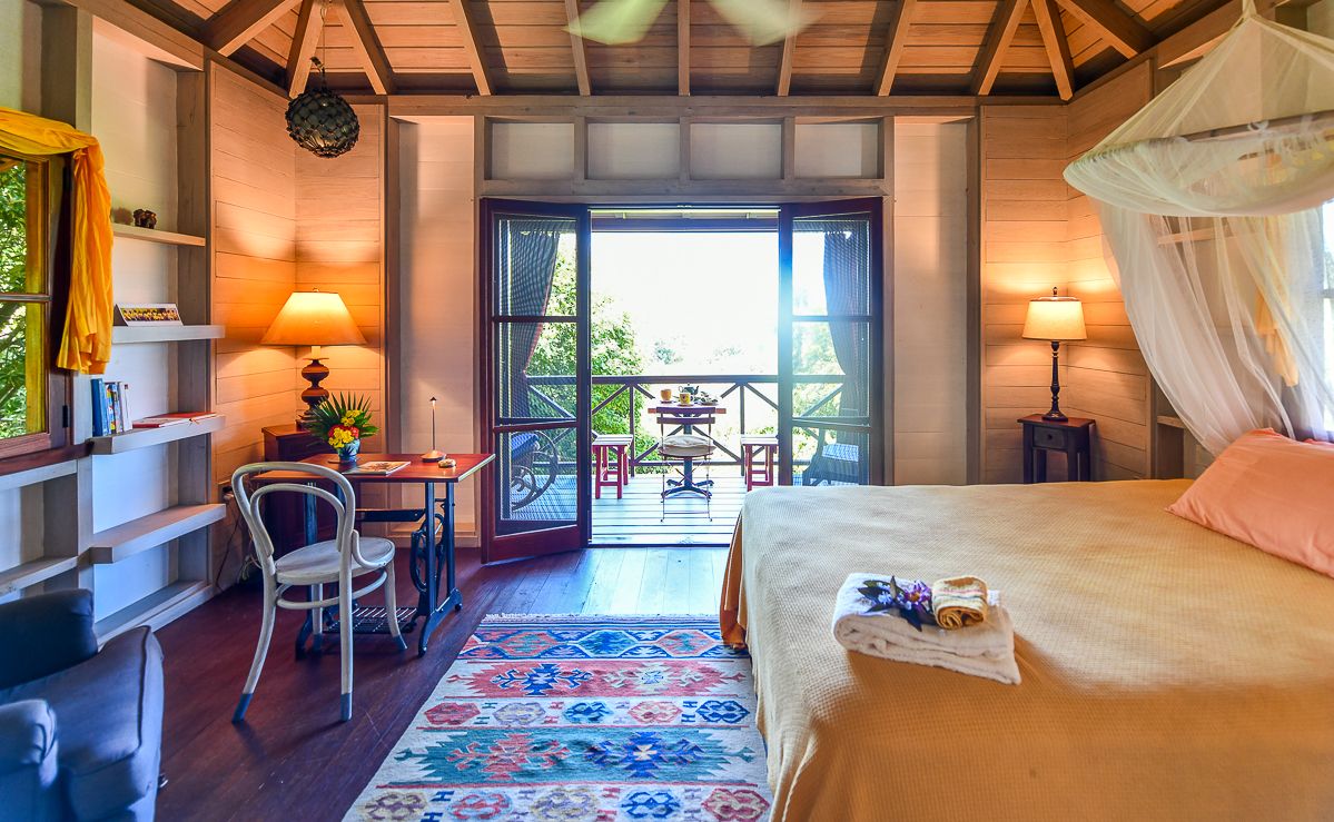 Cozy tropical bungalow bedroom with canopy bed, colorful rug, wooden walls, and a balcony overlooking lush greenery.
