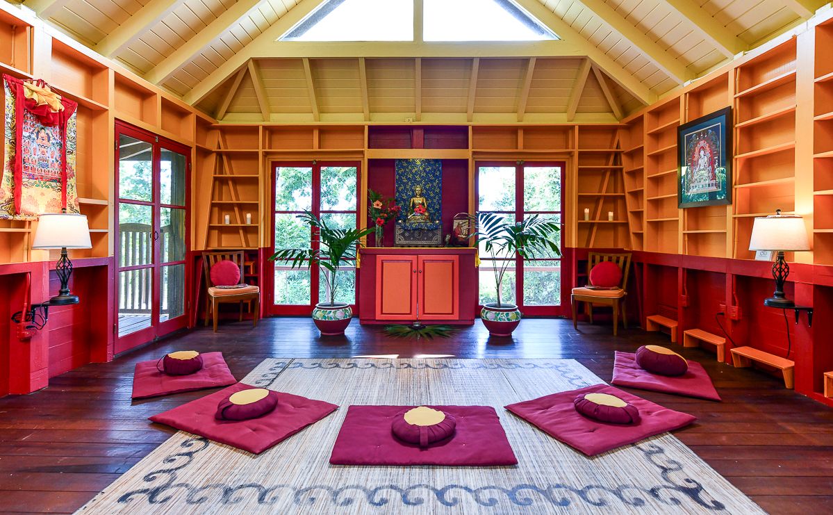 Peaceful meditation room with red decor, Buddha shrine, floor cushions, and wooden bookshelves in yoga retreat.