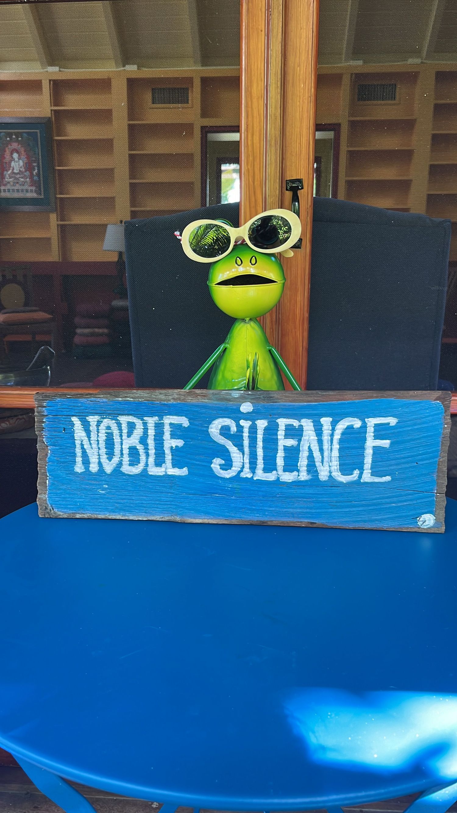 A green frog figurine wearing sunglasses sits behind a blue wooden Noble Silence sign on a bright blue table.