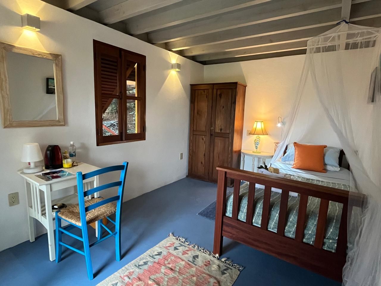 Cozy hotel room with canopy bed, blue chair, wooden desk, rustic wardrobe, and colorful rug on blue floor.
