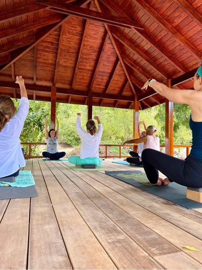 Group of women practicing seated yoga with raised arms in a wooden open-air pavilion surrounded by tropical greenery.