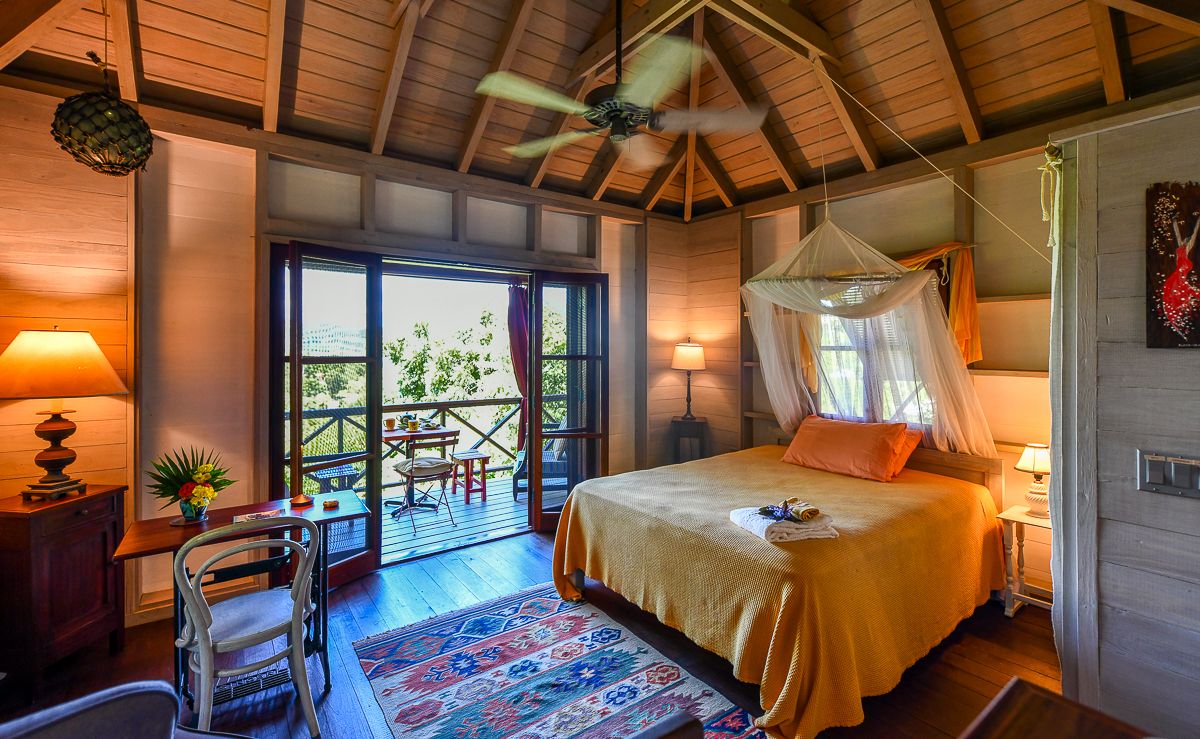 Tropical bungalow bedroom with canopy bed, vaulted wood ceiling, ceiling fan, orange accents, and balcony with lush garden views.