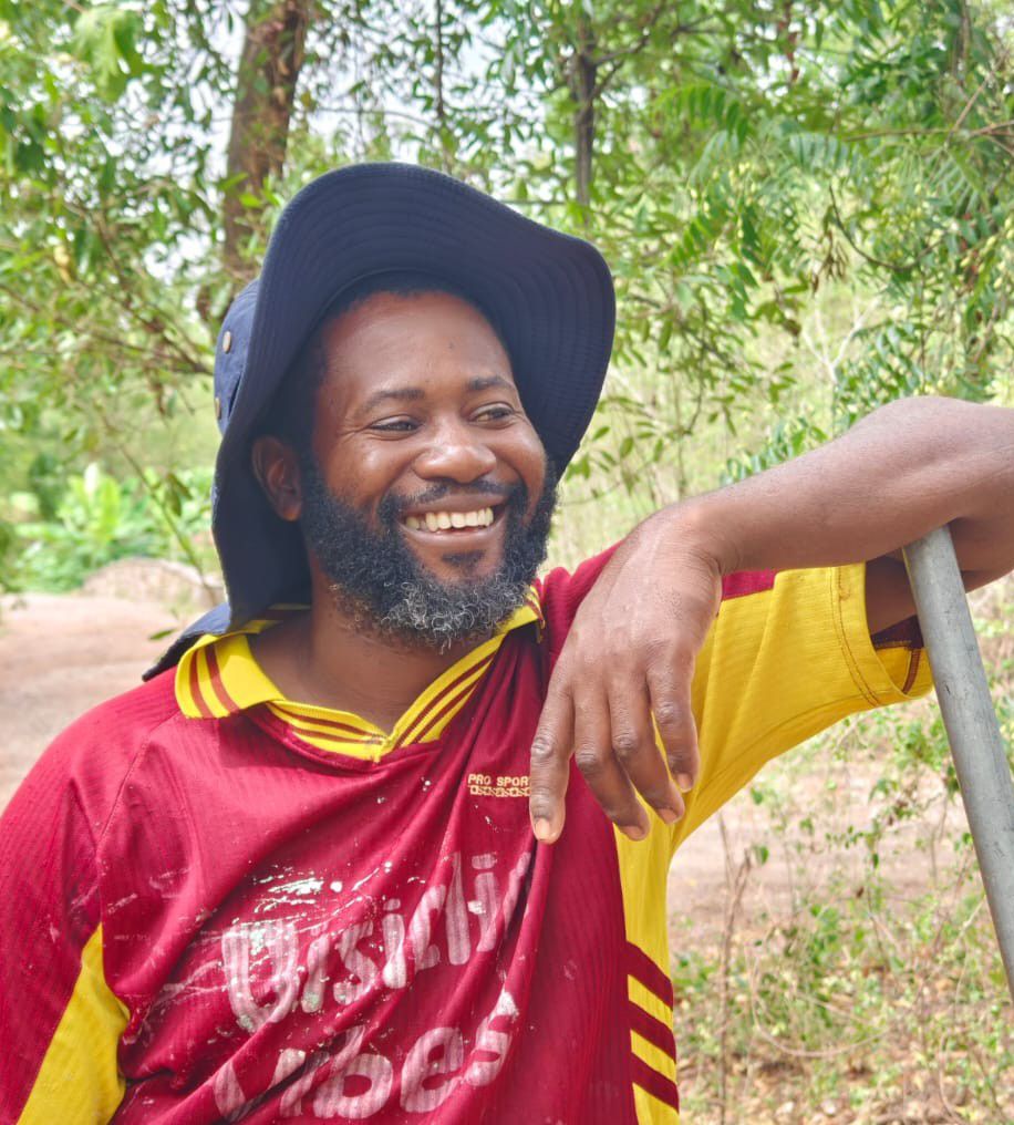 Smiling bearded man wearing a blue bucket hat and red yellow jersey, leaning on a stick outdoors.