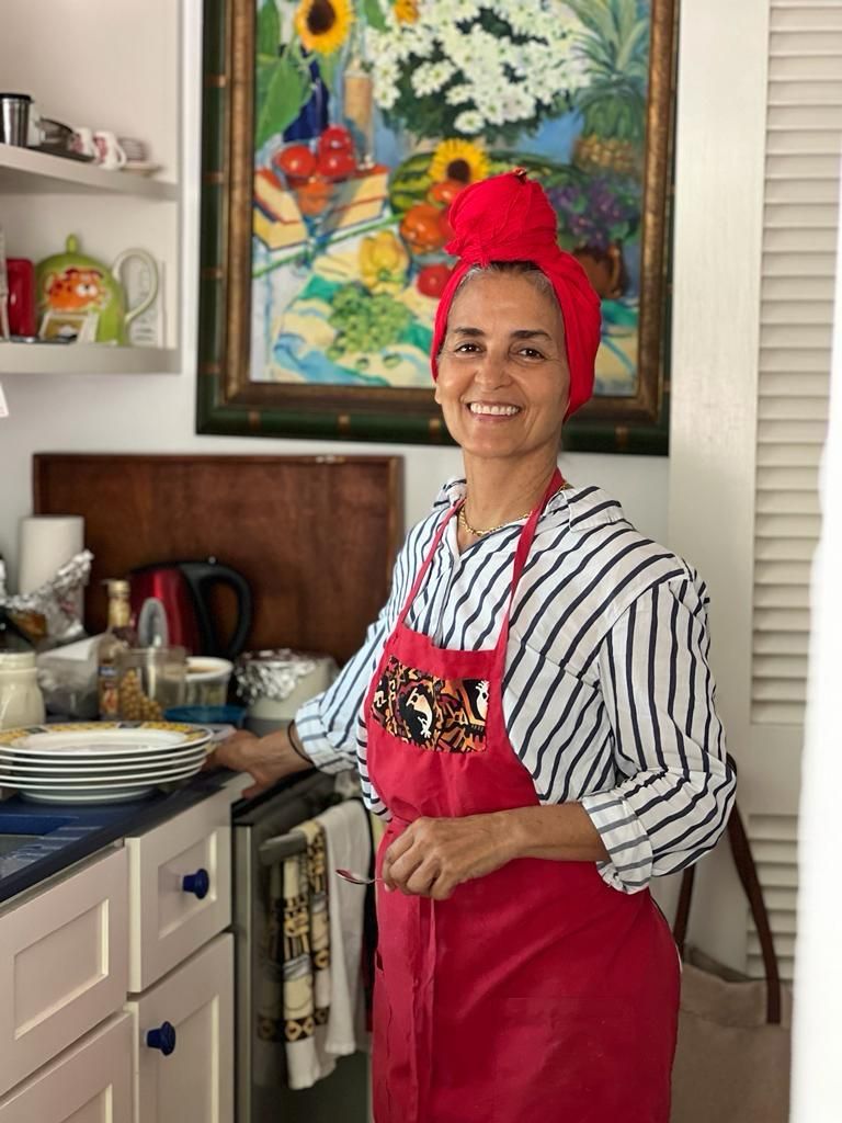 Woman in red apron and head wrap smiling while cooking in a colorful kitchen with vibrant artwork.