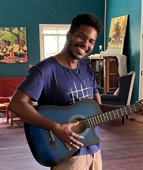 Smiling man holding a blue acoustic guitar in a colorful room with artwork on teal walls.