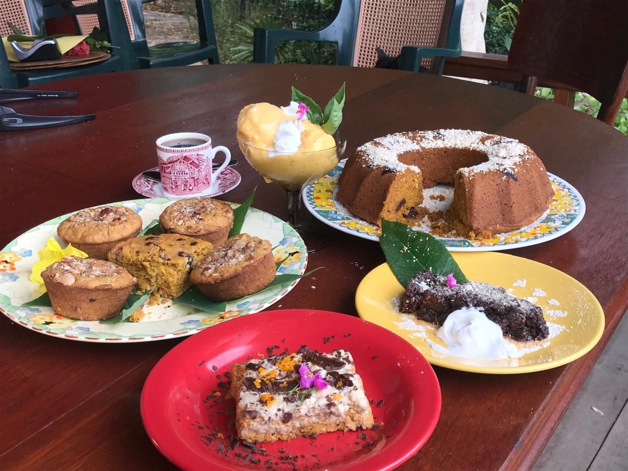 Assorted homemade cakes and muffins displayed on colorful plates on a wooden table, including a bundt cake dusted with powdered sugar.