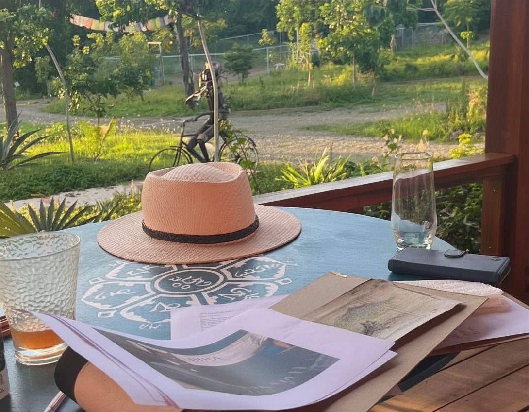 A pink panama hat rests on a glass table with books and drinks, overlooking a lush tropical garden at sunset.